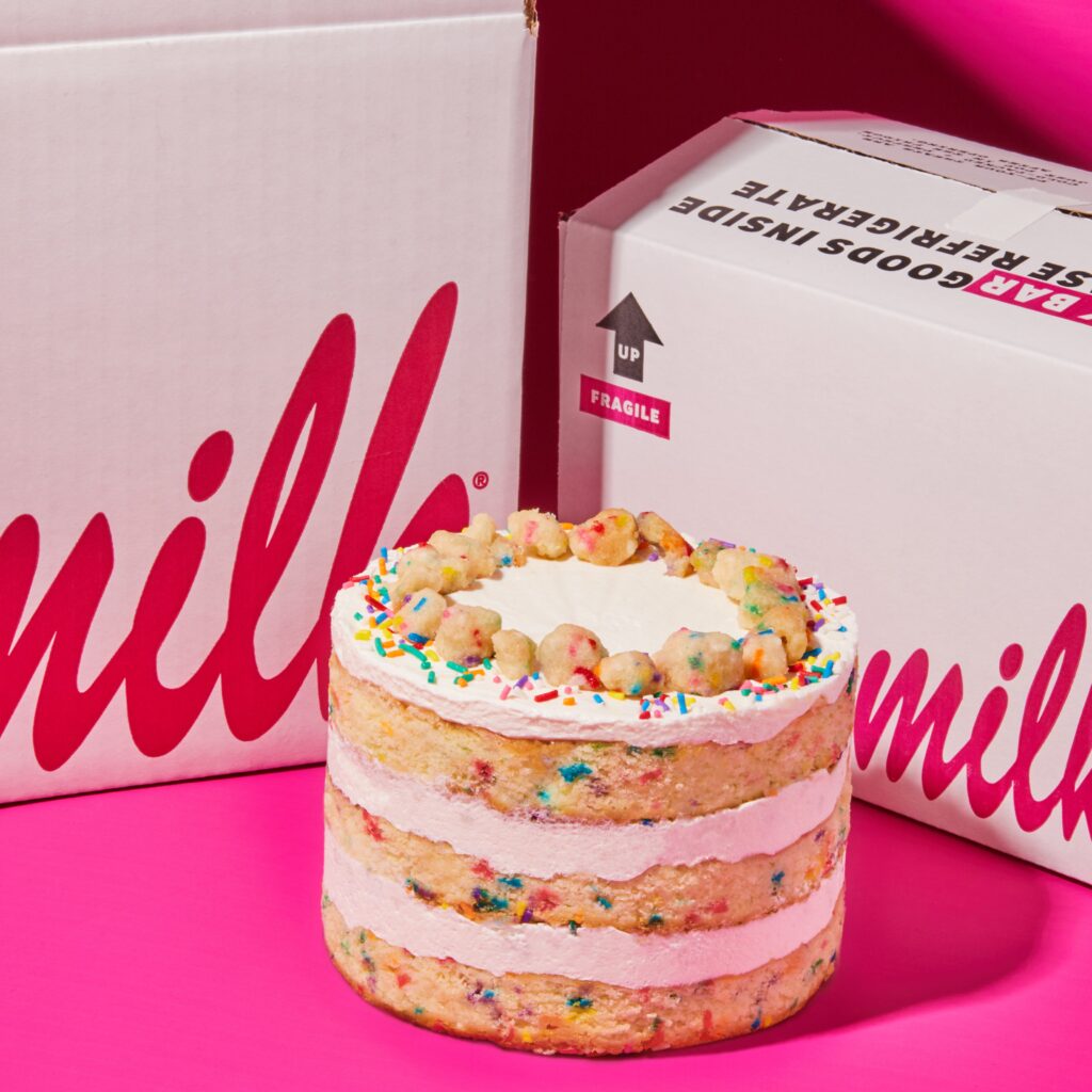 A three-layer Milk Bar cake with frosting and sprinkles sits in front of two white boxes against a pink background. The cake is elegantly decorated with colorful sprinkles and crumbles on top. Claim your free Milk Bar cake today and savor the delightful experience!
