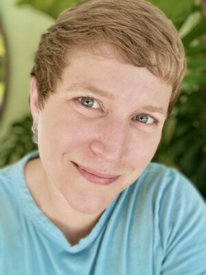 A person with short light brown hair and blue eyes smiles gently, wearing a light blue shirt. Lush green leaves blur in the background, giving this Employee Spotlight a bright and natural feel perfect for Pride 2025.