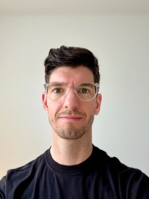 A man with short dark hair, clear-framed glasses, and a trimmed beard is wearing a black shirt and standing in front of a plain, light-colored background for an LGBT+ Employee Spotlight.