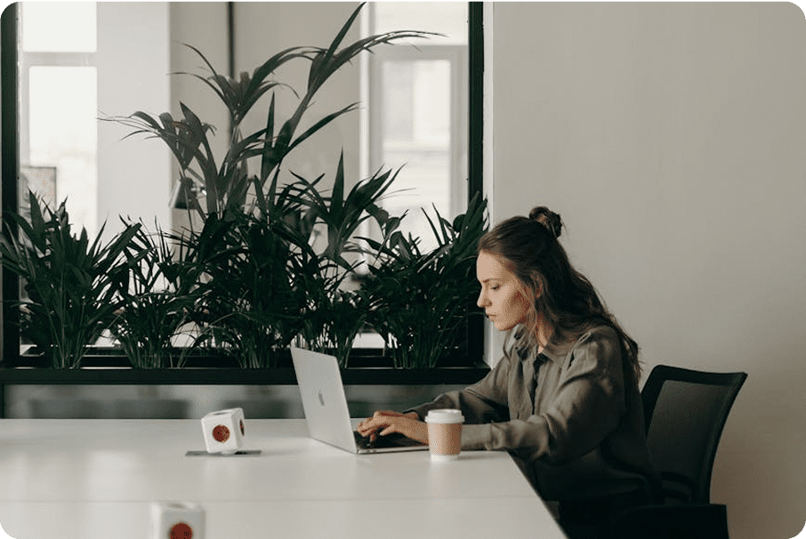 A woman sits at a desk working on a laptop in a modern office with potted plants in the background, analyzing Net Revenue Retention. A coffee cup and a mug are on the white table next to her.