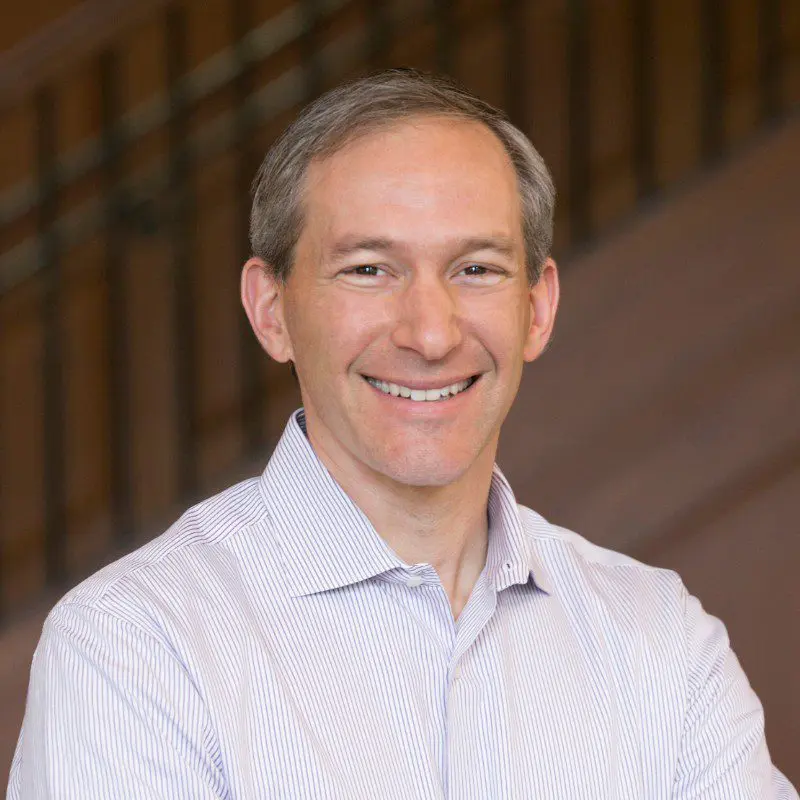 A middle-aged man with short gray hair smiles while wearing a light striped dress shirt. He is standing indoors in front of a blurred staircase railing, ready to transform customer experience at scale.
