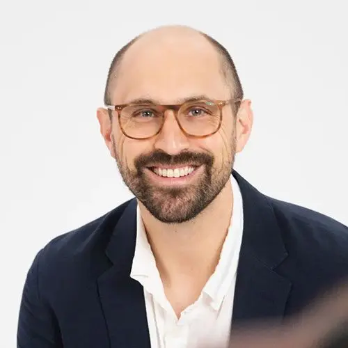 Josh Schachter, a smiling man with a beard, wearing glasses, a dark blazer, and a white shirt, poses against a plain white background.
