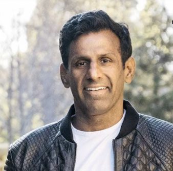 Nick Mehta, a man with short dark hair, wearing a black quilted jacket over a white shirt, smiles outdoors with blurred trees in the background.