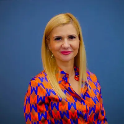 A woman with straight blonde hair wearing a vibrant orange and blue patterned blouse stands in front of a plain blue background, smiling at the camera.