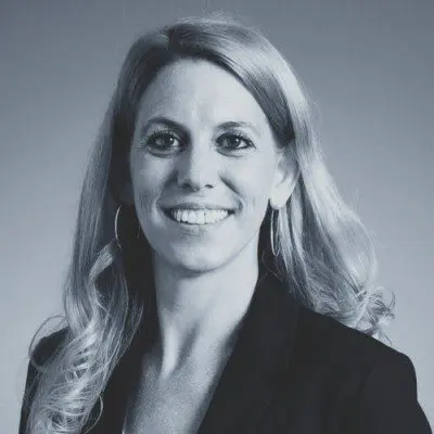 A woman with long, wavy, light-colored hair, wearing a dark blazer and hoop earrings, smiles at the camera against a plain background. The image is in black and white.
