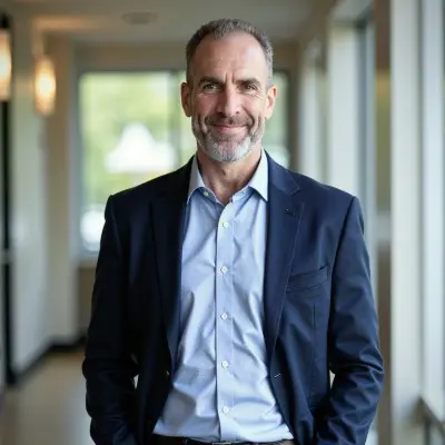 A middle-aged man with short gray hair and a beard, wearing a navy blazer and light blue shirt, smiles while standing in a well-lit hallway with large windows in the background.