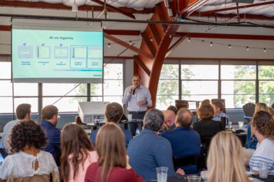 A man speaks into a microphone in front of a seated audience at a presentation. Behind him, a slide titled “AI vs Agents” is displayed, featuring graphics and text from Agentic Connect, in a bright, industrial-style room.