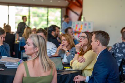 A group of people sit around tables in a bright room, engaged in discussion. Some look attentive, with laptops and notebooks open. The background shows large windows and an Agentic Connect AI Leader near colorful sticky notes.