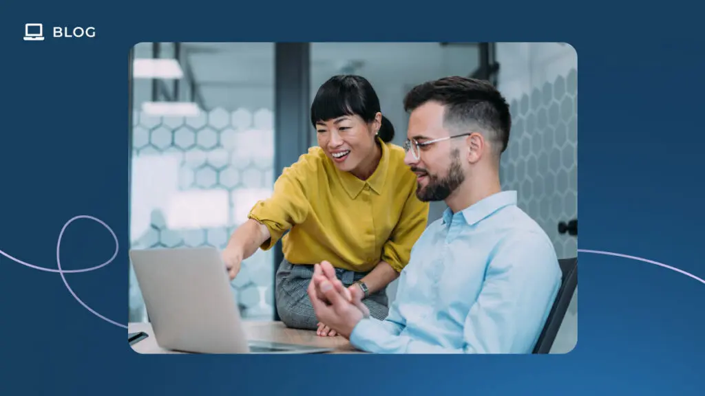 Two people in a modern office sit at a desk with a laptop. A woman in a yellow shirt is smiling and pointing at the screen, discussing the best time to send an NPS survey, while a man in glasses and a blue shirt looks at the laptop attentively.