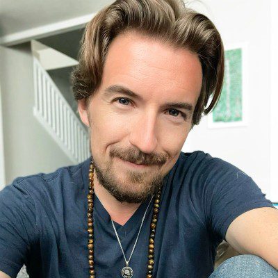 A man with light brown hair and a beard smiles at the camera. He is wearing a dark t-shirt, a beaded necklace, and a pendant. A staircase and framed artwork are visible in the bright background.