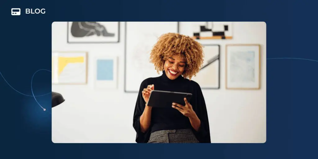 A smiling woman with curly hair sits indoors, holding a stylus and using a tablet, as she explores AI customer engagement solutions, with framed artwork on the wall behind her.