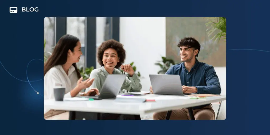 Two people stand indoors, discussing something while looking at a laptop. One gestures with their hands as the other holds the device, reflecting a spirit of community. Green plants are visible in the background.