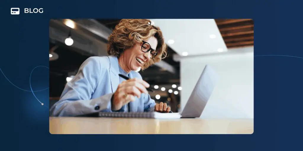 A smiling woman with curly hair and glasses sits at a desk, looking at a laptop and writing in a notebook, inspired by the latest Unchurned Podcast about using AI at work as her superpowered sidekick in a modern office setting.