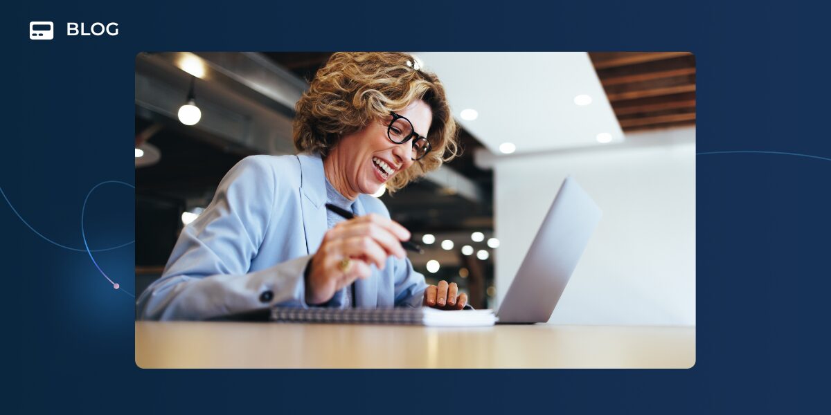 A smiling woman with curly hair and glasses sits at a desk, looking at a laptop and writing in a notebook, inspired by the latest Unchurned Podcast about using AI at work as her superpowered sidekick in a modern office setting.