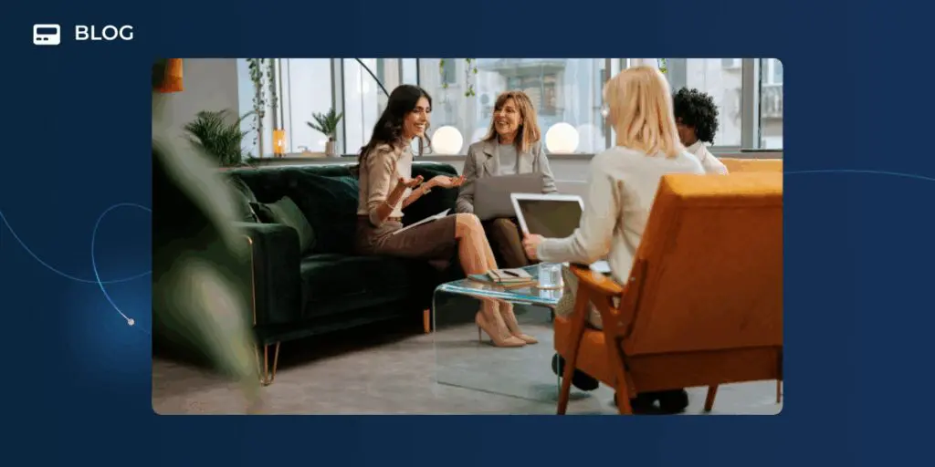 Four people sit in a modern office, engaged in a lively conversation about the Successful Super User Program. Two women on a dark green sofa face two others seated in armchairs, with a glass coffee table between them. The setting is bright and professional.