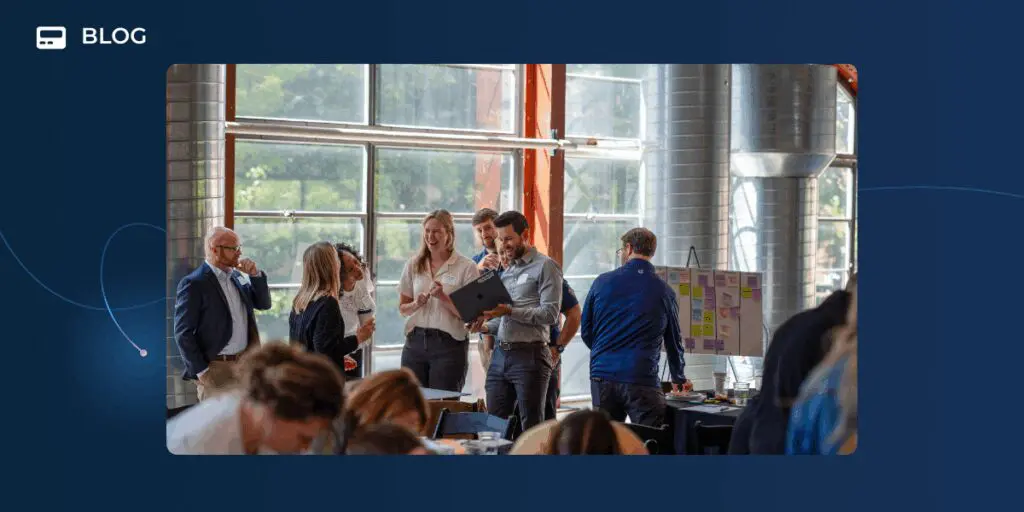 A group of people stand and talk around a laptop in a bright, modern office space with large windows and industrial decor; others, discussing Agentic Connect, are working at tables in the foreground.