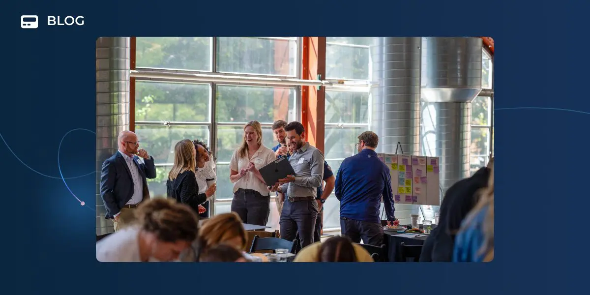A group of people stand and talk around a laptop in a bright, modern office space with large windows and industrial decor; others, discussing Agentic Connect, are working at tables in the foreground.