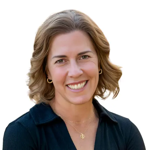 Erica Kuhl, a woman with shoulder-length light brown hair, wearing a black top, small gold hoop earrings, and a gold necklace, smiles at the camera against a white background.