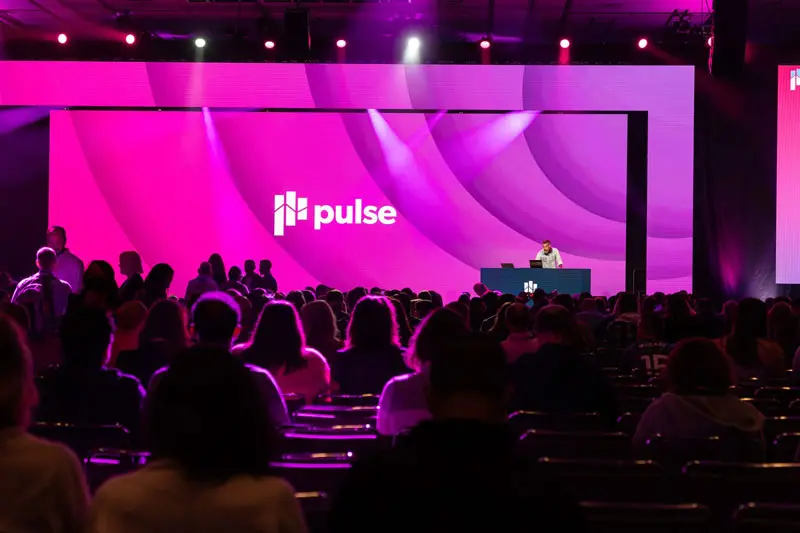A large audience sits facing a stage with bright pink and purple lighting, where a Customer Success speaker stands at a podium in front of a screen displaying the word "pulse" and a geometric logo.