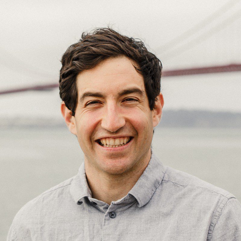 A man with short dark hair smiles at the camera, wearing a light gray shirt. In the blurred background, there is water and a bridge structure on a cloudy day—this is ex-Google PM Jacob Bank, now innovating in AI Agents.