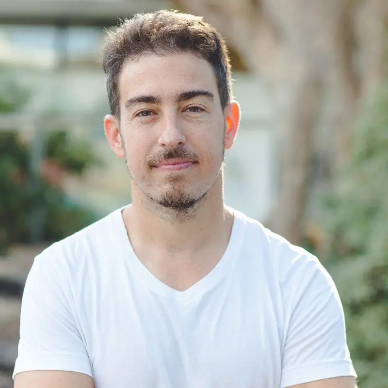 A man with short brown hair and a trimmed beard is standing outdoors, wearing a white t-shirt. He is looking at the camera with a slight smile, with blurred greenery and trees in the background, embodying the confidence of top CEOs.