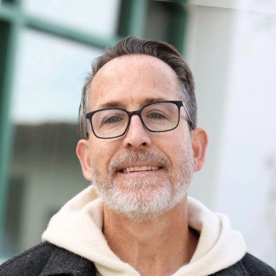 A middle-aged man with short gray hair and a neatly trimmed beard is wearing glasses, a white hoodie, and a dark jacket, smiling outdoors in front of a blurred building with green window frames—perhaps after an ARR meeting at Bonfire Ventures.
