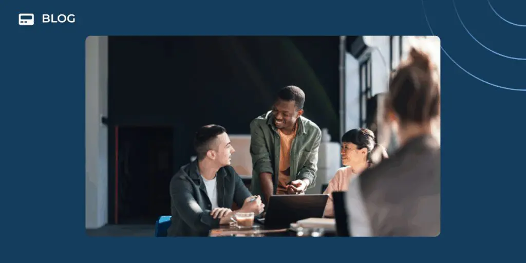 Three people sit at a table with a laptop, smiling and discussing ideas, while another person is blurred in the foreground. The modern office setting reflects how scalable customer communities thrive in collaborative environments.