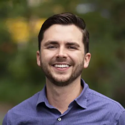 A young man with short brown hair and a beard, smiling and wearing a purple button-up shirt, stands outdoors with blurred greenery in the background.