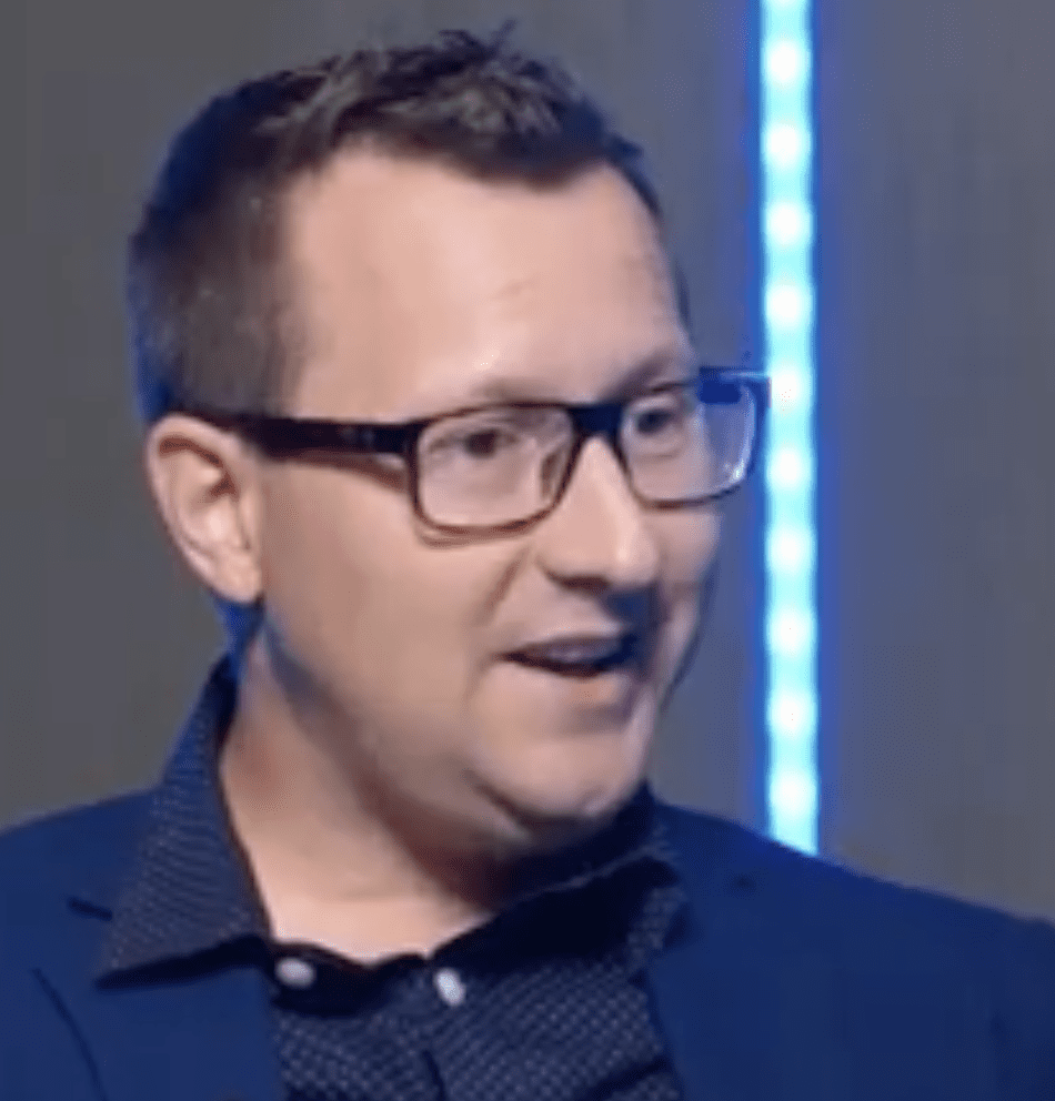 A man with short brown hair and glasses, wearing a dark blue blazer and a patterned shirt, is speaking about AI. The background features a vertical blue light strip.