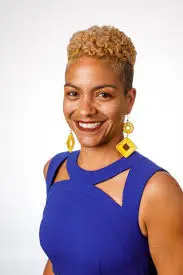 A woman with short curly blonde hair, wearing a sleeveless royal blue dress with cut-out details and yellow geometric earrings, smiles against a plain white background.