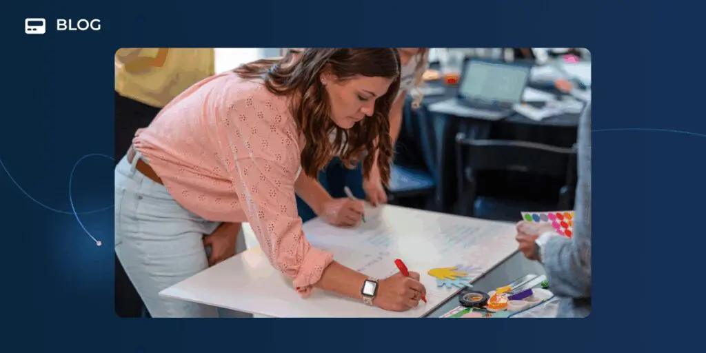 A woman in a pink blouse writes on a large sheet of paper at a table, surrounded by art supplies and others, during a collaborative workshop focused on creative podcast app ideas and gamified podcast promotion.