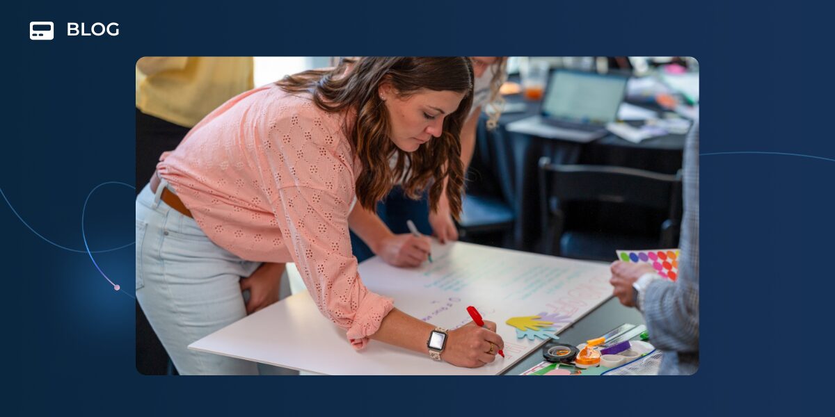 A woman in a pink blouse writes on a large sheet of paper at a table, surrounded by art supplies and others, during a collaborative workshop focused on creative podcast app ideas and gamified podcast promotion.