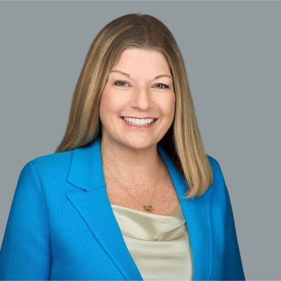 A woman with straight, shoulder-length blonde hair is smiling at the camera. She is wearing a bright blue blazer over a light blouse with layered necklaces—an inspiring image of today’s CS Leaders in Customer Success. The background is a plain gray.