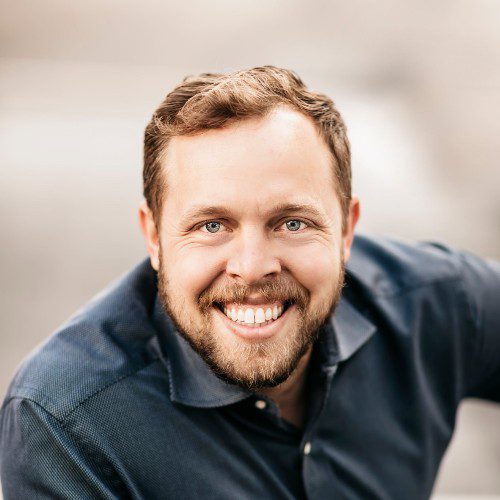 A man with light brown hair and a beard, wearing a dark blue collared shirt, smiles broadly at the camera against a blurred, neutral background—reflecting the energy of leading small team AI workflows.