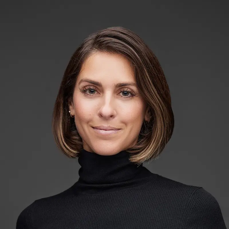 A woman with straight, shoulder-length brown hair and light makeup is wearing a black turtleneck sweater, posing in front of a plain dark background and smiling softly at the camera, reflecting a welcoming community spirit.