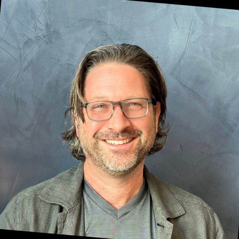 A man with shoulder-length brown hair, a beard, and glasses is smiling. Wearing a gray t-shirt and dark jacket, he stands in front of a textured gray background—ready to reveal the biggest GTM mistake (spoiler alert: stop chasing CAC!).