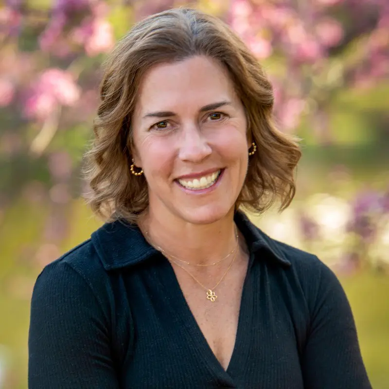 A woman with wavy brown hair smiles while standing outdoors in a garden-like setting, wearing a black top and gold jewelry. The scene suggests she enjoys being part of a supportive community focused on growth and retention.
