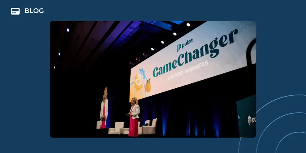 A woman stands on stage at a Pulse event in front of a large screen displaying "2025 GameChangers Award Winners," with two empty chairs beside her and a blue background surrounding the image.