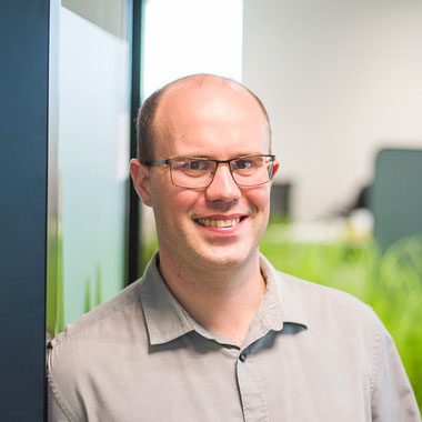 Jef Vanlaer, smiling with glasses and a light gray collared shirt stands indoors, leaning against a glass wall in a modern office.