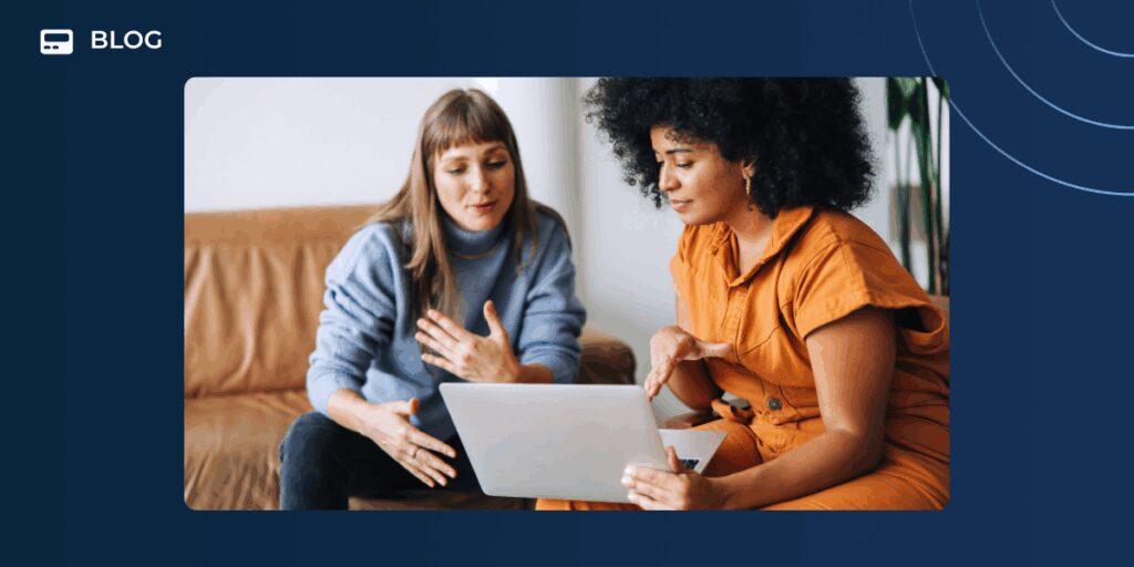 Two women sit on a couch, engaged in discussion while looking at a laptop. One gestures as they collaborate on a digital customer success strategy, both appearing focused and engaged. The setting is casual and comfortable.