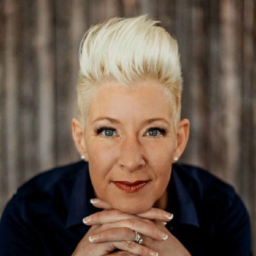 Courtney Hauser, a person with short, platinum blonde hair styled upward smiles softly, resting their chin on folded hands with manicured nails, wearing a dark collared shirt, and sitting in front of a blurred wooden background.