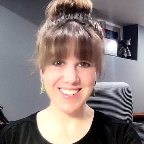 Photo of Jeni Asaba with light brown hair in a bun and bangs smiles at the camera. She is wearing green earrings and a black top, sitting in a room with gray walls.