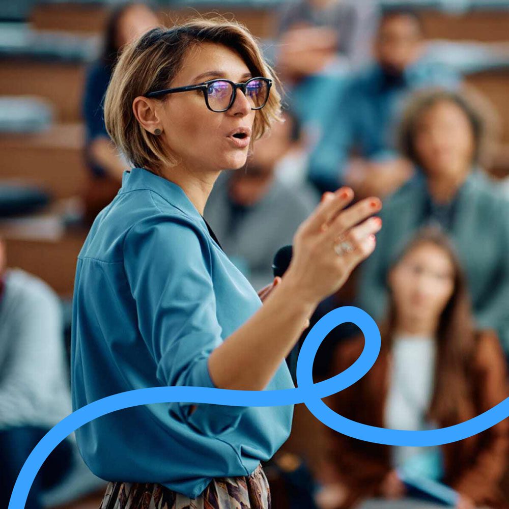 A woman wearing glasses and a blue shirt speaks into a microphone in front of an audience in a lecture hall. A blue squiggly line is overlaid at the bottom of the image.