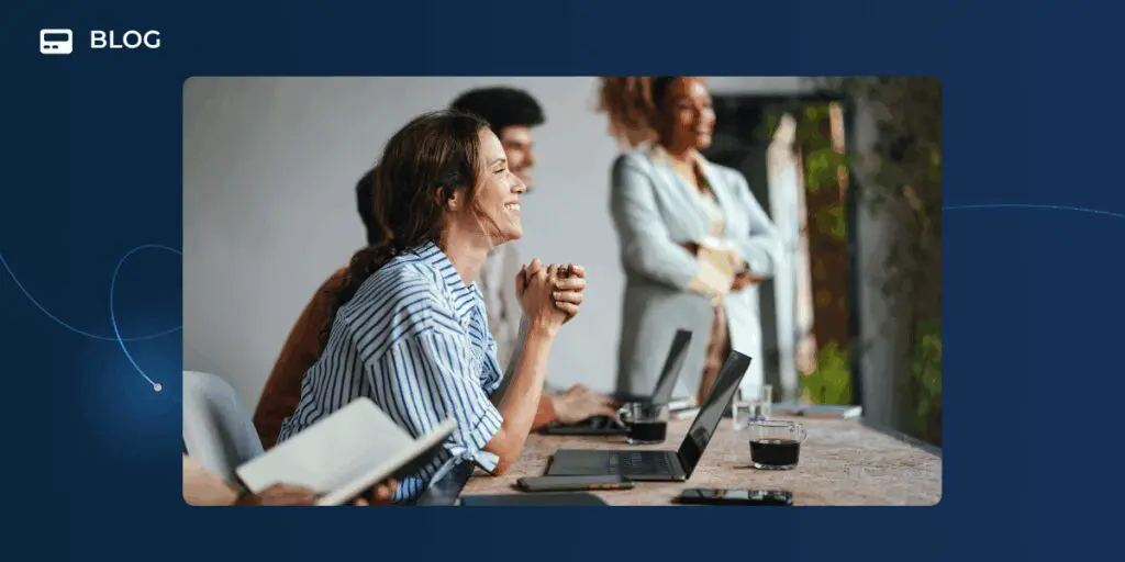 A group of people sits at a desk with laptops and notepads, engaged and smiling, while a woman stands in the background. The casual, collaborative work environment suggests a team discussing artificial intelligence and AI ethics.