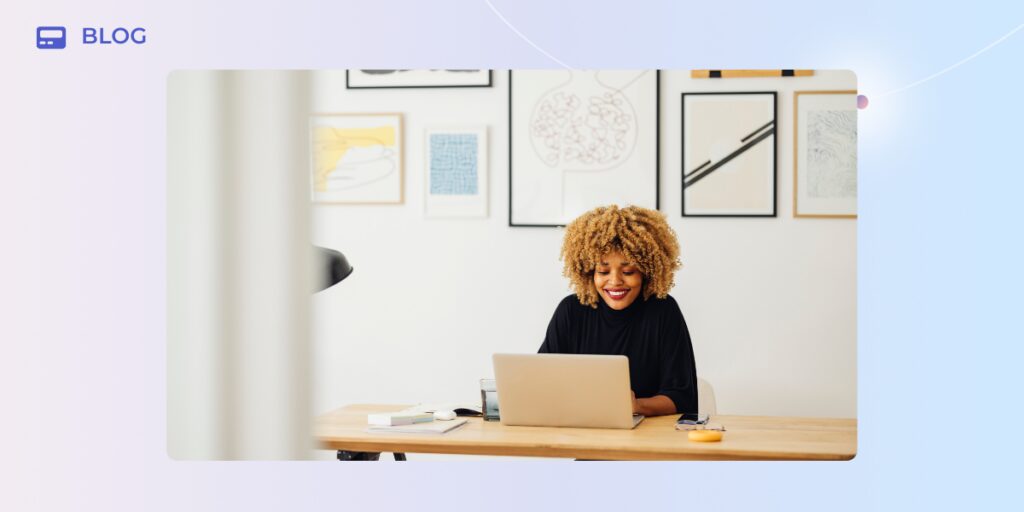 A woman with curly hair sits at a desk, smiling at her laptop. The modern, bright space features framed artwork and a "Blog" label. Perfect for SaaS experts sharing insights on AI SaaS retention strategies.