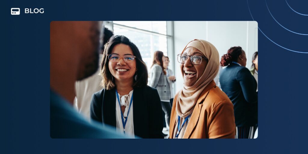 Two professionally dressed women are smiling and chatting about AI at a networking event, with others mingling in the background. The image features a blue border and "BLOG" text in the top left corner, highlighting the importance of staying human at scale.