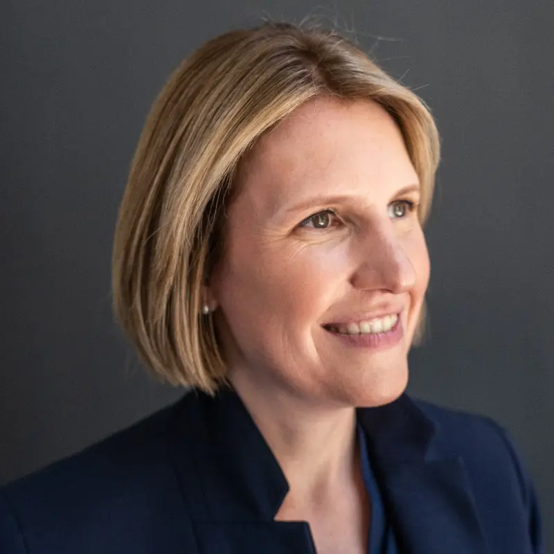 A woman with short blonde hair, wearing a navy blazer, smiles while looking slightly to the right against a plain dark background, embodying the confidence often seen in leaders of top AI Companies.