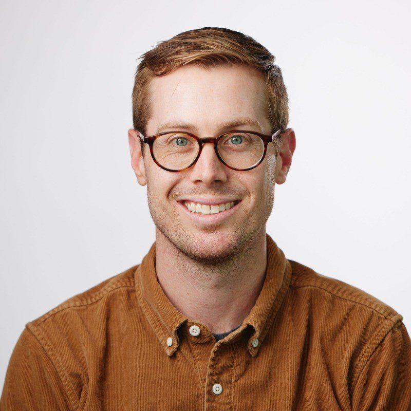 A man with short light brown hair, wearing round eyeglasses and a brown corduroy button-up shirt, smiles at the camera against a plain light background—perfect for profiles at innovative AI companies.