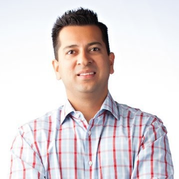 Abbas Haider Ali, with short dark hair and a light-colored, button-up shirt featuring red and blue plaid stripes, stands with arms crossed and smiling against a plain light background, ready to discuss revenue or post-sales budget strategies.