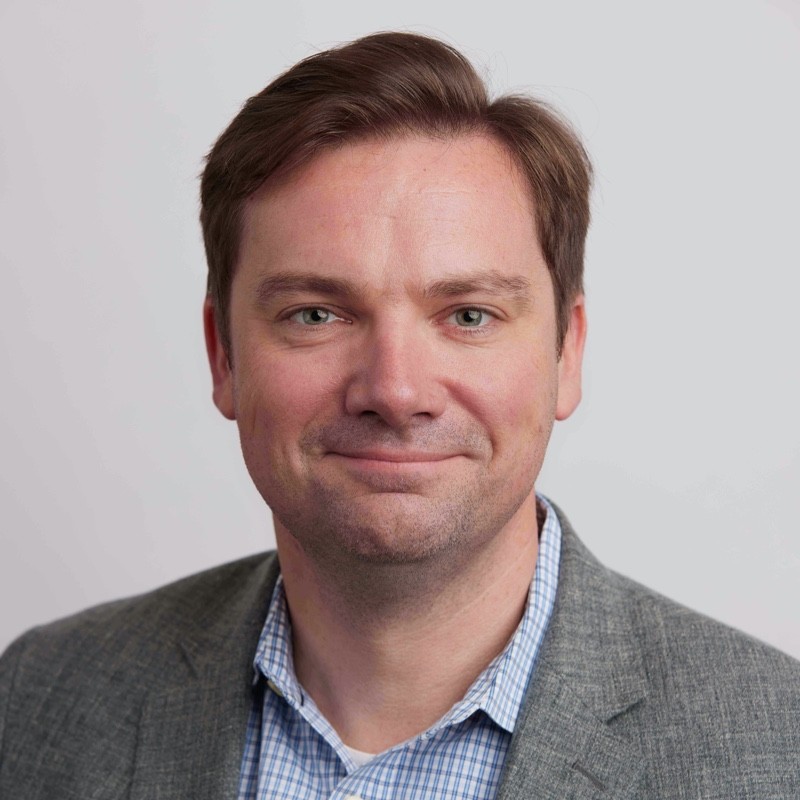 Jim Richmond, CCO at Smartling, smiles slightly. He is wearing a gray blazer over a blue and white checked shirt, posing in front of a plain light gray background.